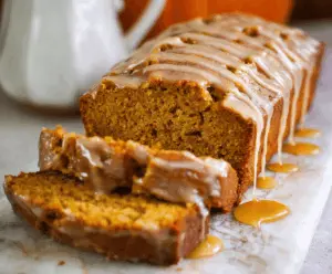 Delicious homemade pumpkin bread topped with a glossy maple glaze, served on a rustic wooden platter with cinnamon sticks and a cup of coffee.